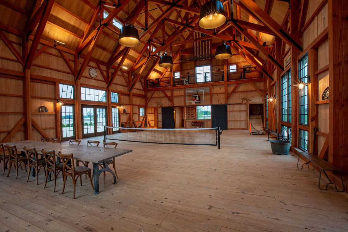 A spacious indoor gym in a beautifully converted bank barn with high wooden ceilings, large windows, and exposed beams. A tennis net is set up in the center, a basketball hoop at the end, and a long wooden dining table with chairs sits to the side.