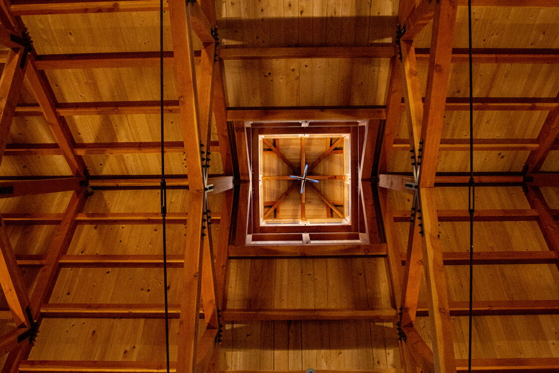 A symmetrical view looking up at a wooden ceiling with exposed beams and two large round chandeliers, featuring warm lighting and a central skylight—evoking the craftsmanship of a luxury horse barn or an elegant bank barn.