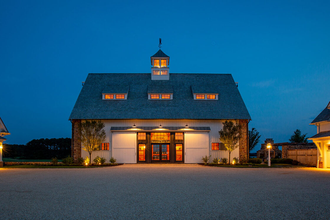 A large, well-lit party barn with a cupola and multiple windows glows warmly at dusk, flanked by two smaller buildings under a deep blue evening sky.
