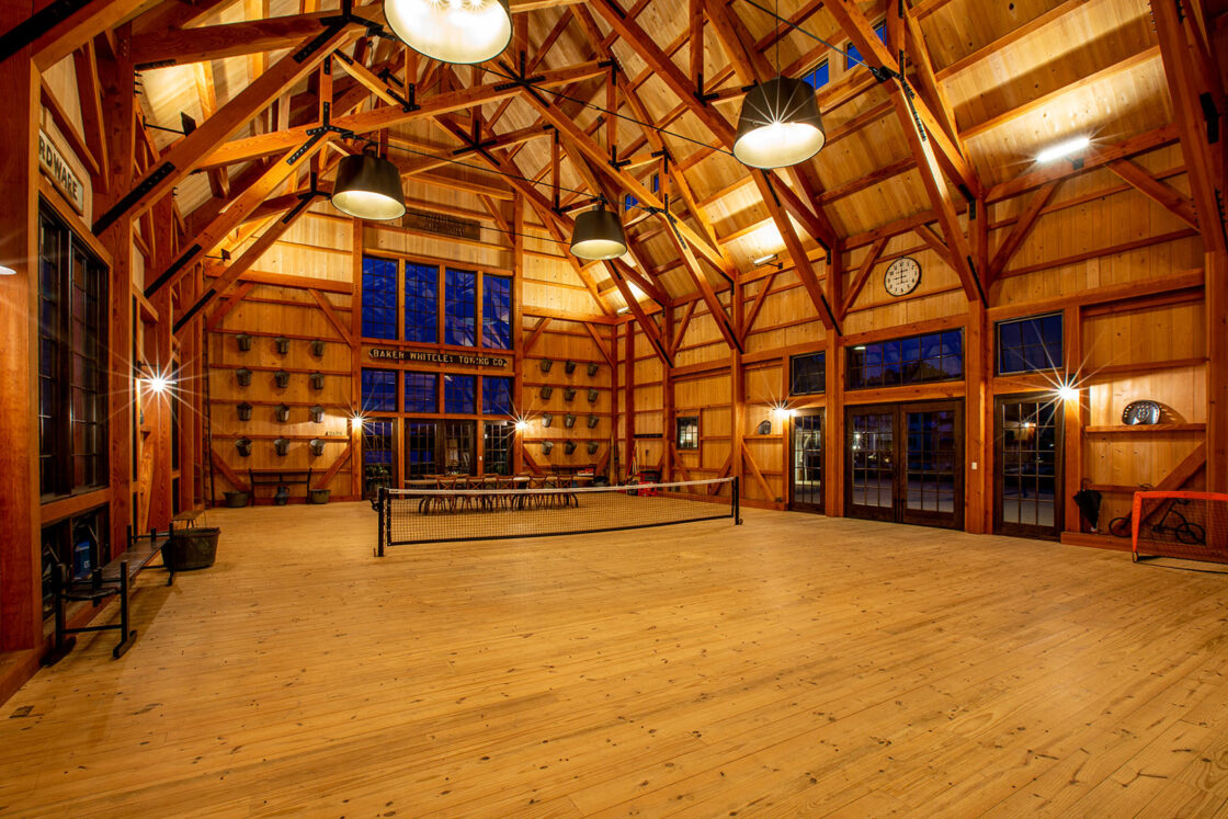 A spacious indoor party barn with wooden beams and floors, large windows, and bright overhead lights. A ping-pong table and chairs are set up in the center, with animal mounts and a large clock on the walls.
