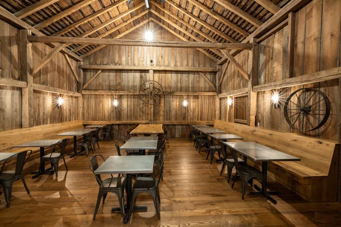 A rustic dining area with wooden walls, ceiling, and floor evokes a luxury horse barn vibe. Metal chairs and wooden tables line the space, while wagon wheels and warm lights add to the cozy, party barn atmosphere.