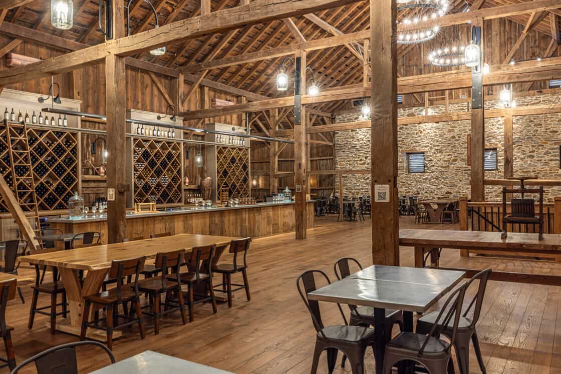 A spacious rustic winery tasting room, reminiscent of a luxury home, features wooden beams, high ceilings, wine racks, chandeliers, stone walls, and wooden tables with black chairs arranged throughout the open area.