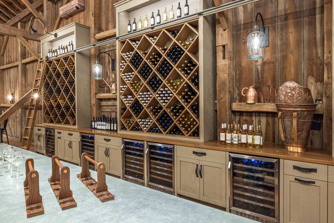 A rustic wine cellar with wooden shelves filled with wine bottles, glass-front wine coolers below, and a countertop displaying wine and glasses—perfect for a luxury home inspired by the timeless charm of a bank barn, with wooden walls and decorative copper items.