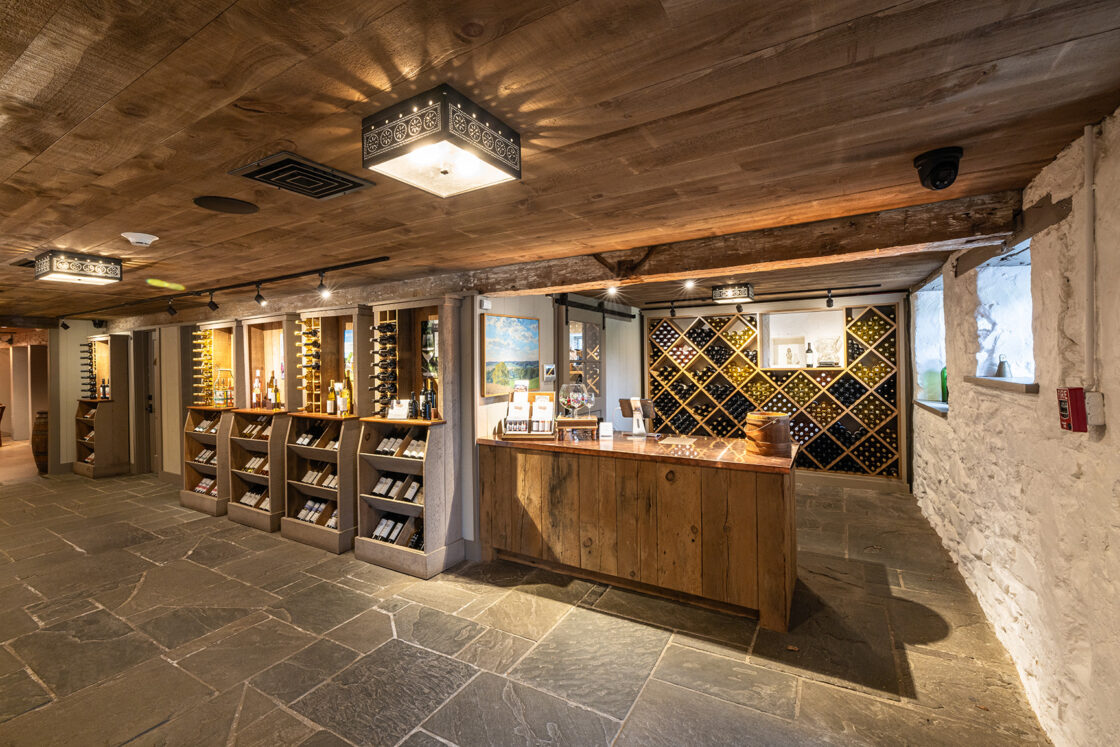 A cozy wine cellar with stone floors, wooden ceiling, shelves of wine bottles, a wooden counter, and a wine rack against the back wall under warm lighting—perfect for a luxury home or party barn.