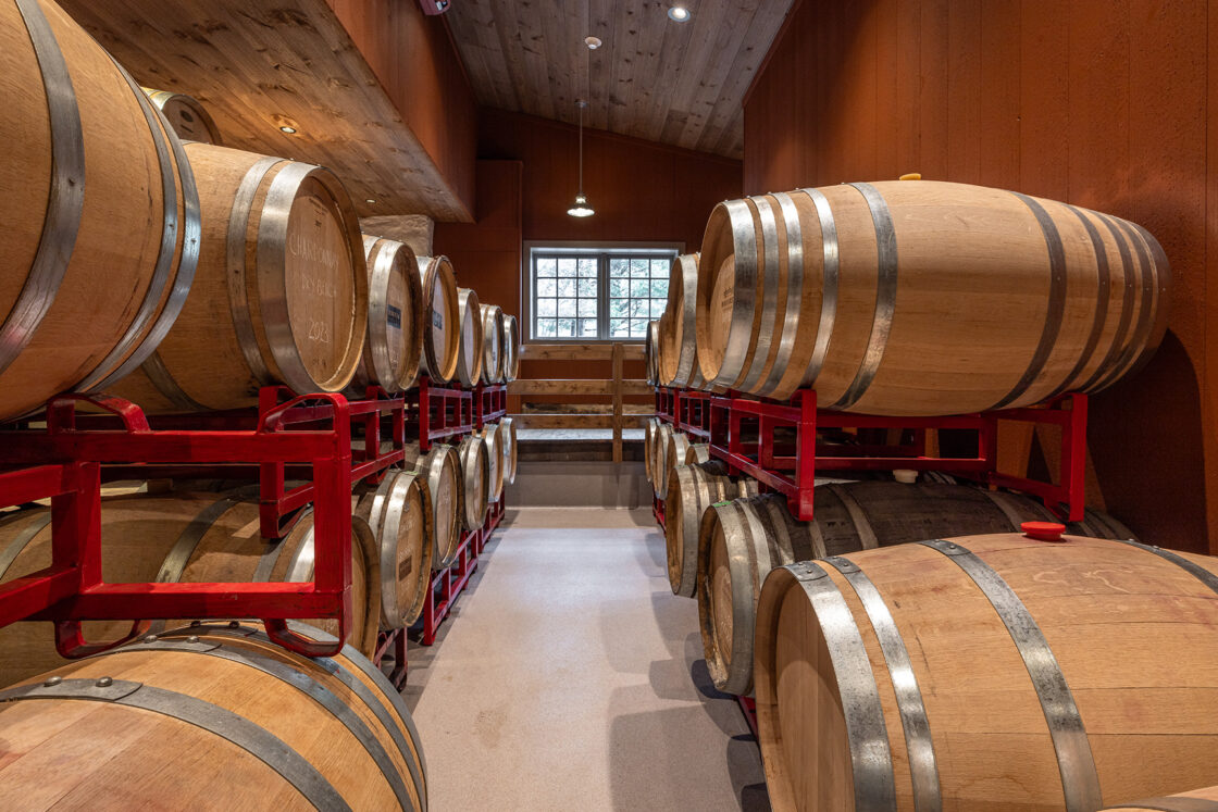 Wooden wine barrels are stacked on red metal racks in a dimly lit cellar with wooden walls and ceiling, reminiscent of a classic bank barn. A window at the far end lets in natural light, illuminating the room.