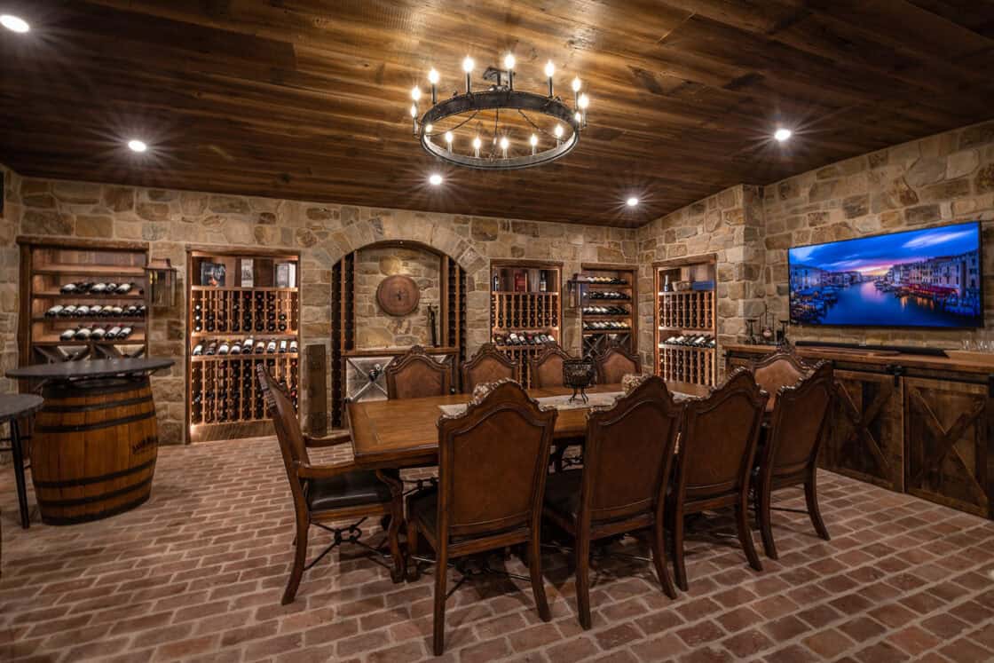 Dining room table with ten chairs surrounding it inside a luxury home’s wine cellar that has a wooden ceiling with a circular light that hangs from it