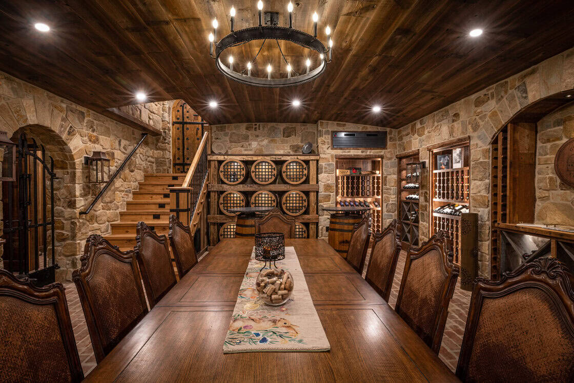 Wooden table sitting in the middle of a luxury home wine cellar that has a wooden ceiling with a circular light fixture all surrounded by stone walls