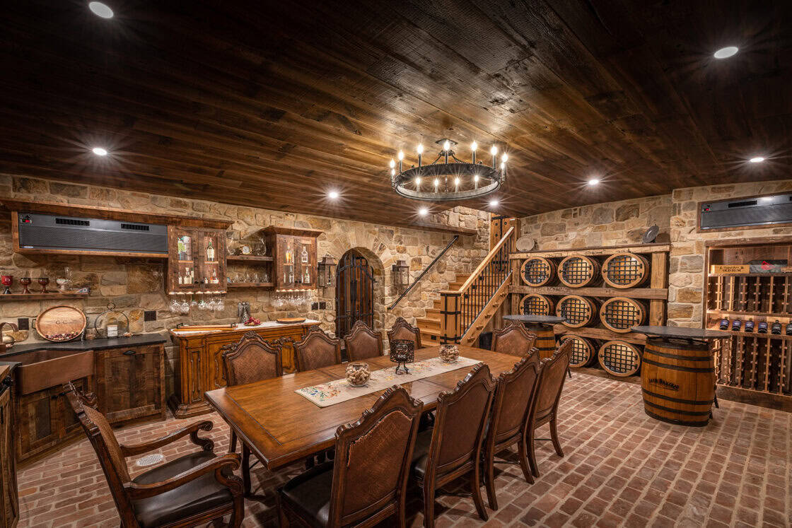 Dining room table with ten chairs surrounding it inside a luxury home’s wine cellar that has a wooden ceiling and two jack daniels barrels made into tables visible in the rear