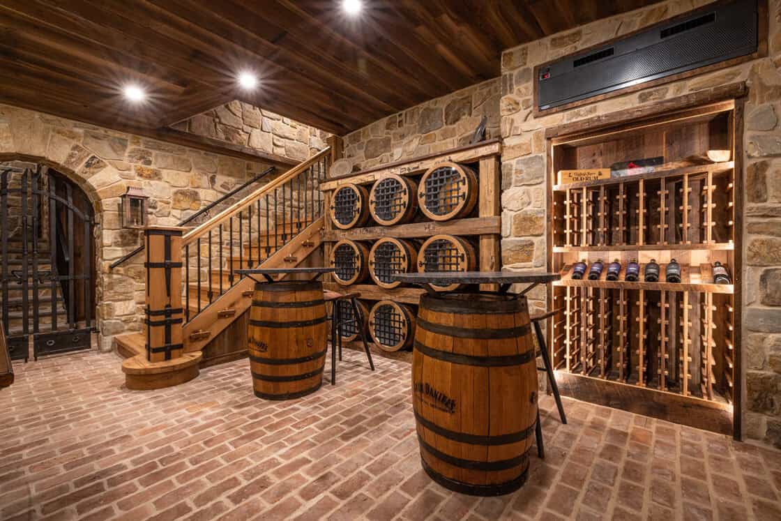 Two jack daniels barrels converted into tables sitting inside a luxury home wine cellar with a wooden ceiling and a brick floor