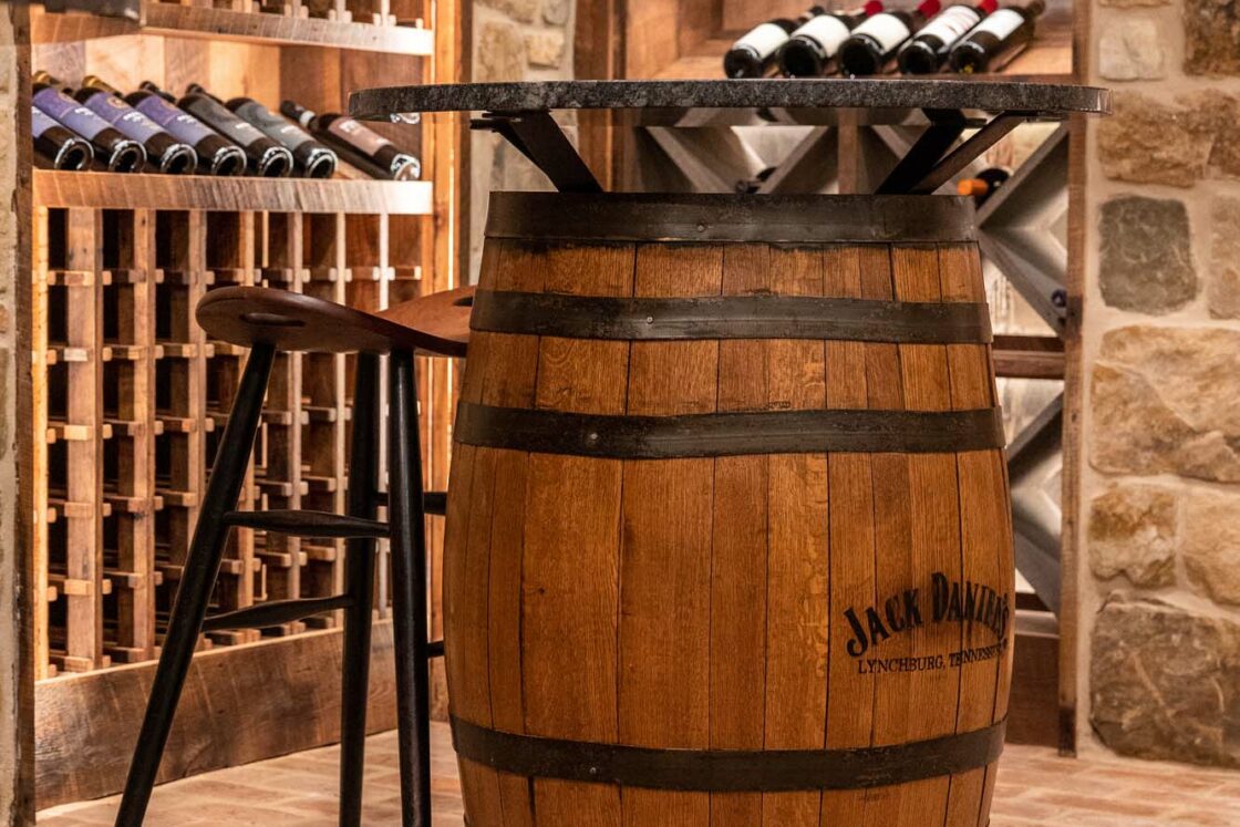 A wooden Jack Daniel’s barrel used as a table with a chair beside it, set in a luxury home wine cellar with bottles of wine stored on wooden racks against a stone wall.