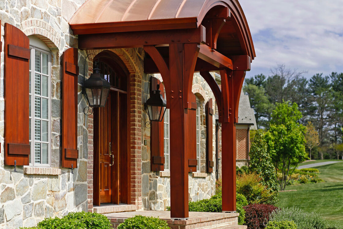 A large stone luxury home with a wooden front door, arched wooden porch with columns, two lantern lights, shuttered windows, landscaped bushes, and a brick path leading to a green lawn on a sunny day.