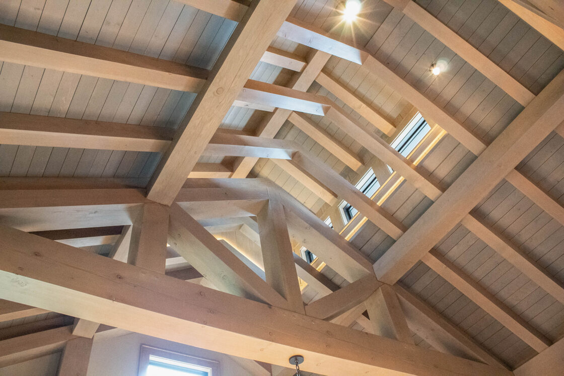 Exposed wooden beams and trusses form a geometric pattern on a vaulted ceiling, perfect for a luxury horse barn or party barn. Skylights and recessed lighting let natural light filter in, highlighting the wood’s texture.