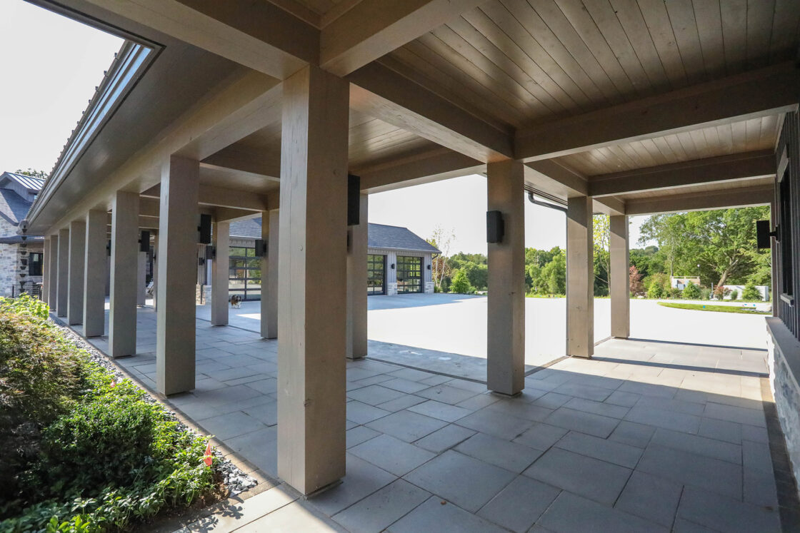 A covered outdoor walkway with stone tile flooring and wooden beams, leading to a sunlit open area reminiscent of a luxury home, with buildings and greenery visible in the background.
