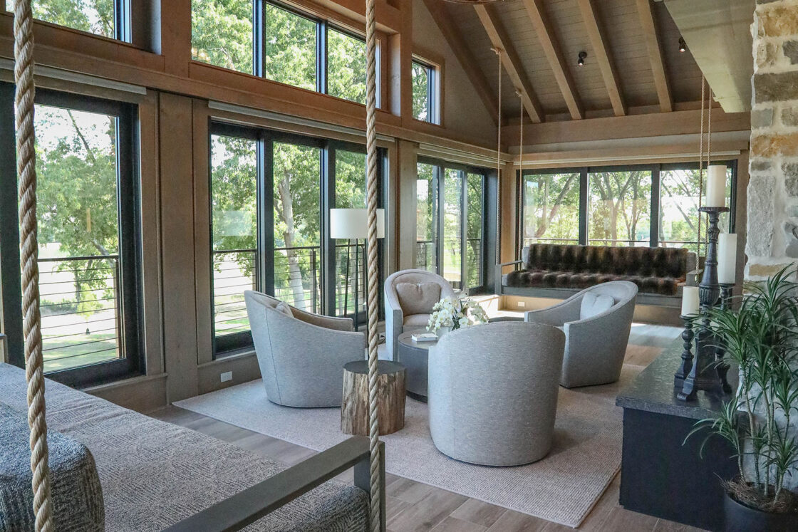 A spacious, modern living room with high wooden ceilings reminiscent of a bank barn, large windows, comfortable seating, a stone fireplace, a rope swing, and abundant natural light. Trees and greenery are visible outside.