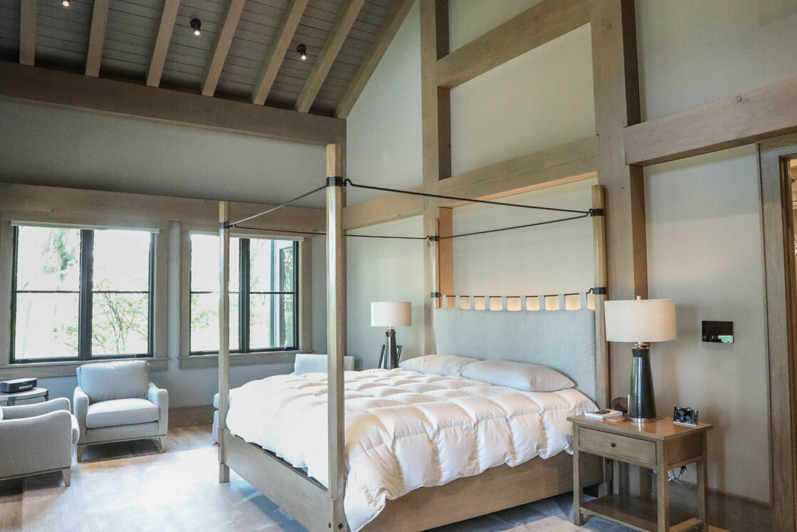 Spacious bedroom with high vaulted wood-beam ceiling reminiscent of a luxury horse barn, large windows, a four-poster bed with white bedding, neutral décor, light wood floors, and modern furnishings. Natural light fills the airy, elegant space.