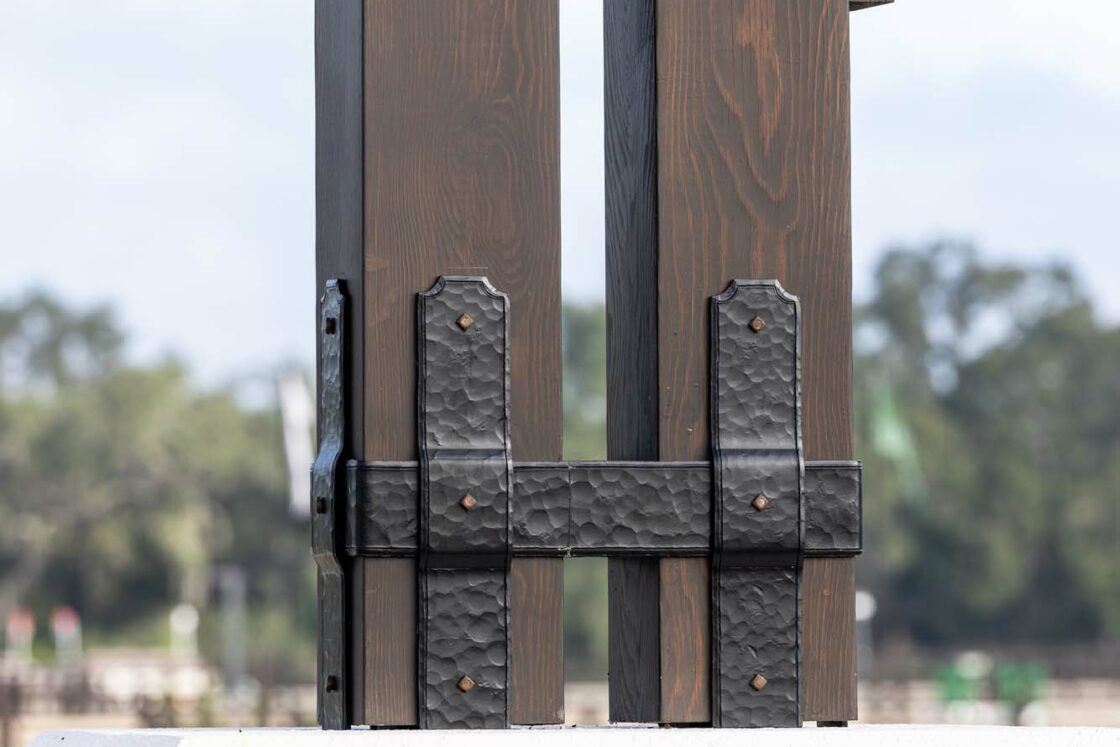A close-up of a large, dark metal hinge attached to vertical wooden beams, which are mounted on a low stone wall—details that add rustic charm to a luxury horse barn. Trees and a cloudy sky are visible in the blurred background.