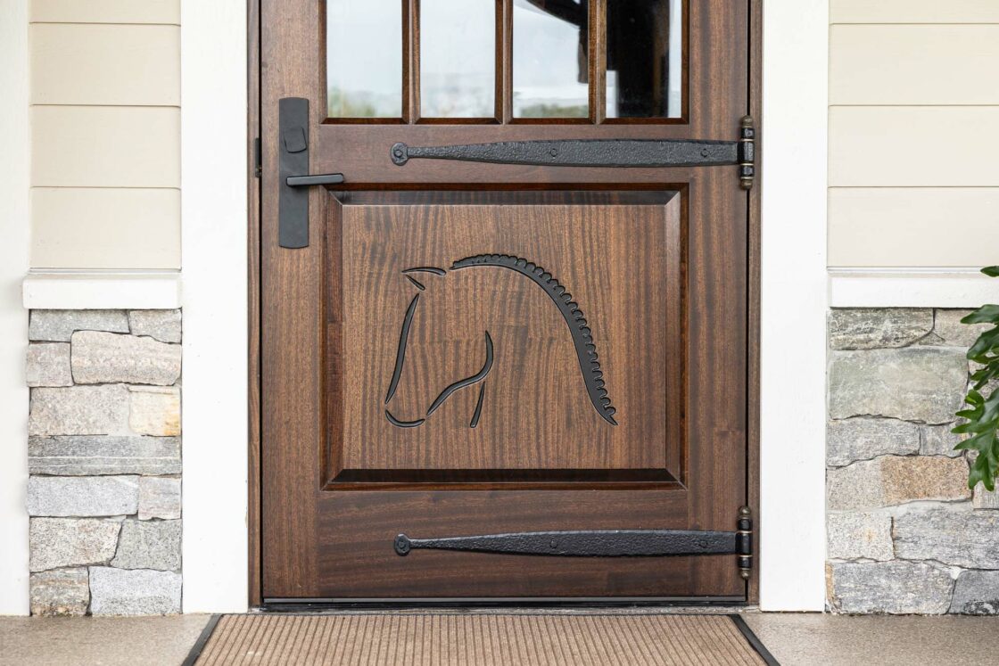 A wooden door with a carved outline of a horse’s head, featuring black iron hinges and handle, evokes the charm of a bank barn. Set in beige siding with a stone base, a doormat lies in front of this inviting entry.