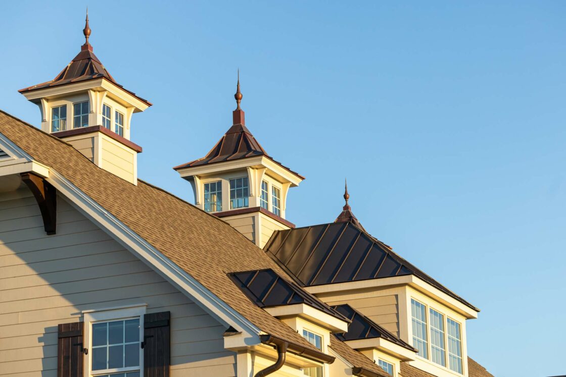 A beige luxury home with brown shingle and metal roofs, featuring three cupolas with windows and decorative finials, is shown against a clear blue sky.