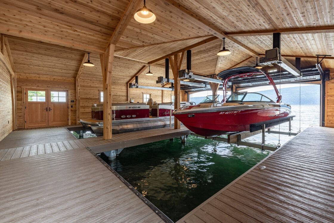 A wooden boathouse with high ceilings features two boats on lifts above the water, with large open doors offering a lake view. The interior, reminiscent of a luxury horse barn, showcases exposed beams and natural wood paneling.