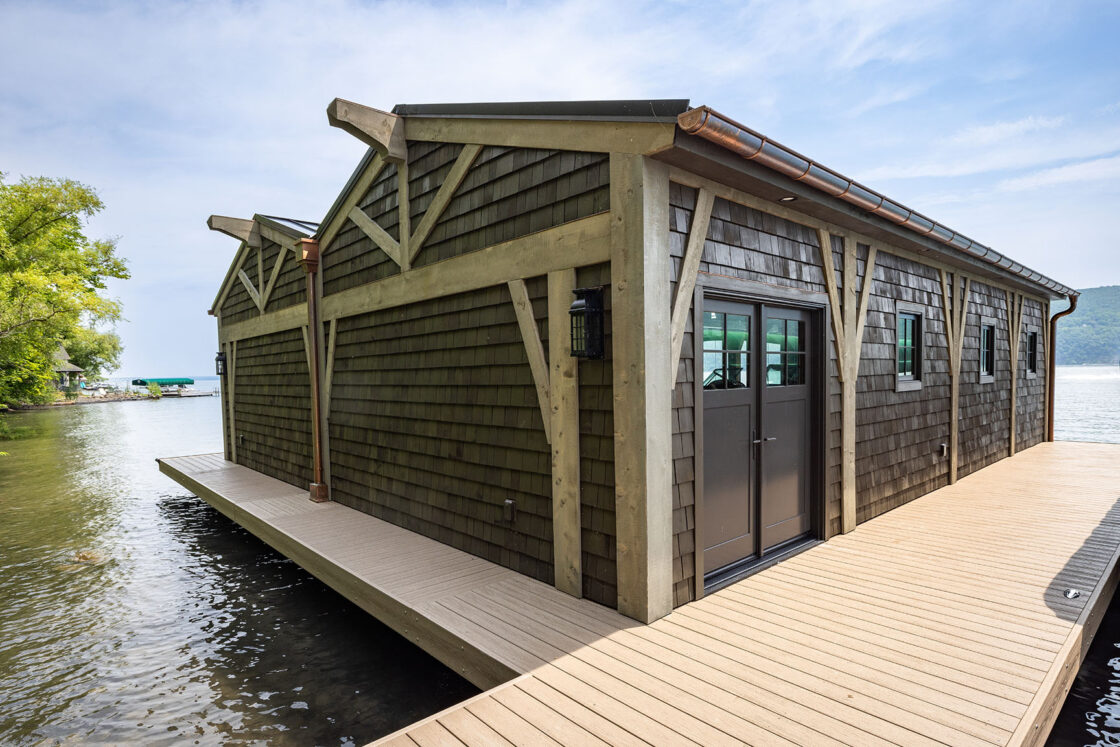 A wooden boathouse with dark shingles and large windows sits on a dock over a calm lake, reminiscent of a luxury home, surrounded by trees and distant hills under a clear blue sky.