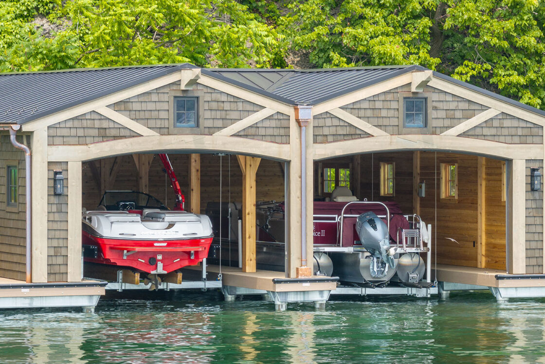 Two covered wooden boat docks sit side by side on the water, each sheltering a boat. One dock holds a white and red speedboat, the other a maroon pontoon boat. Green trees and a nearby luxury home complete the scenic backdrop.