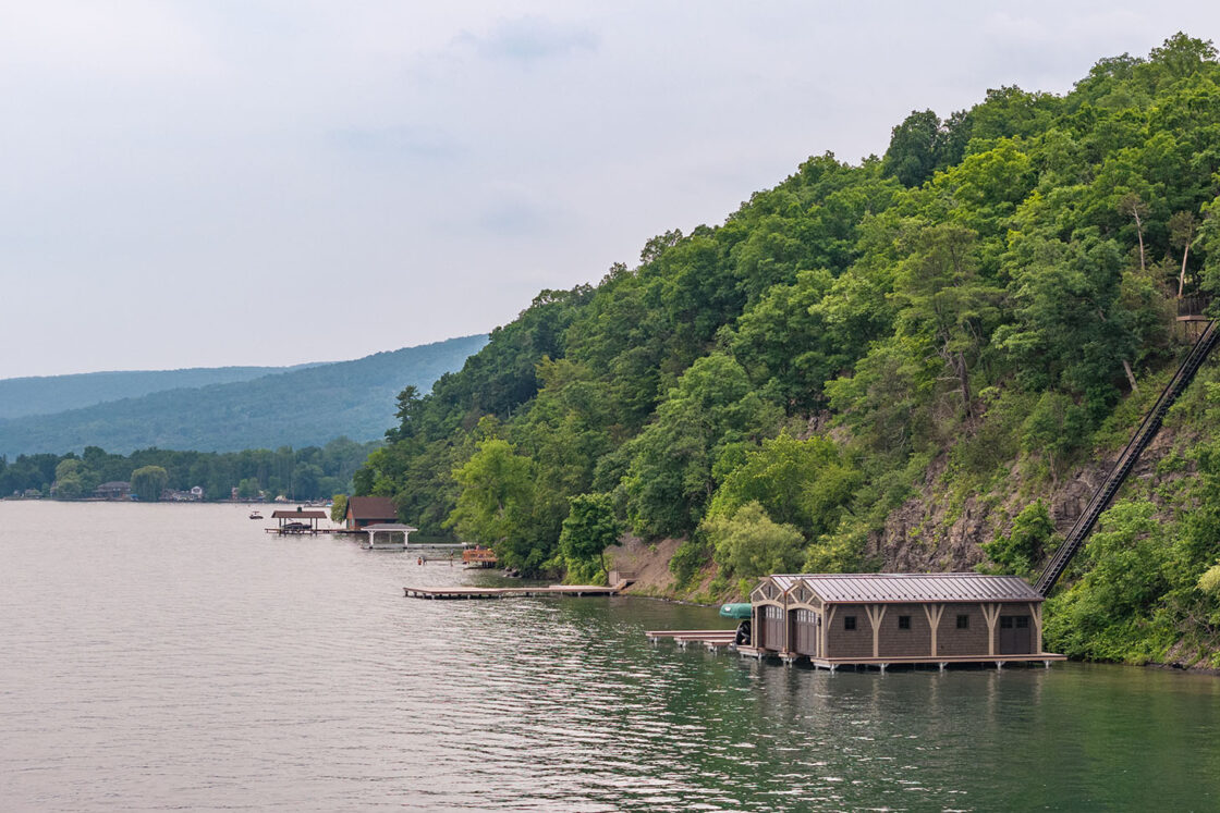 A wooden boathouse sits on the edge of a green, tree-covered hillside along a calm lake, with docks, distant luxury homes, and more houses visible beneath a cloudy sky.