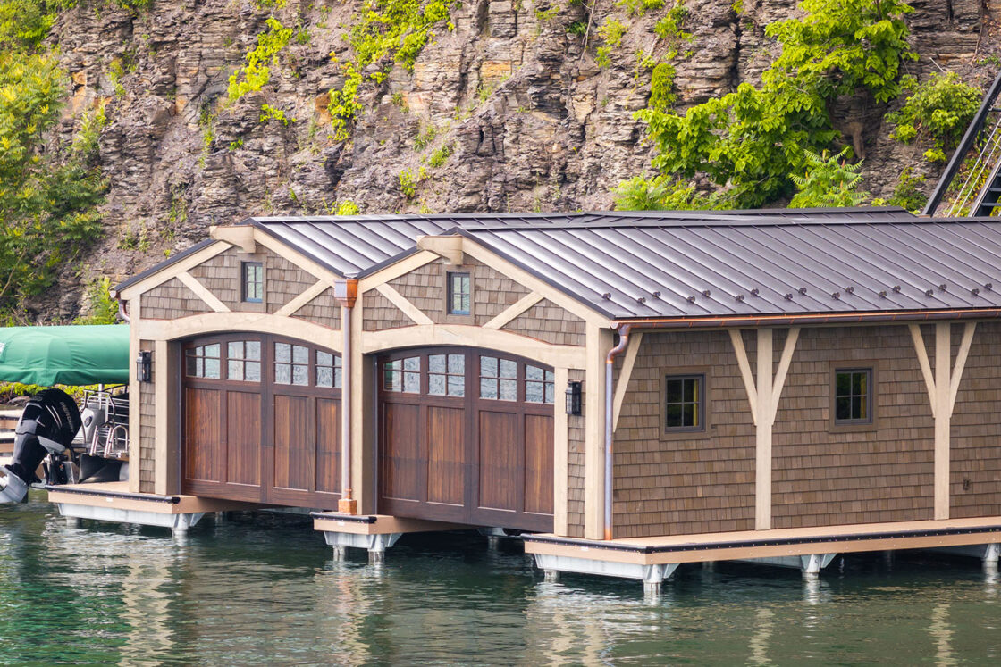 A wooden boathouse with large brown doors sits on a calm lake, backed by a rocky, tree-covered hillside. Reminiscent of a luxury home retreat, an adjacent dock holds a covered boat with an outboard motor.