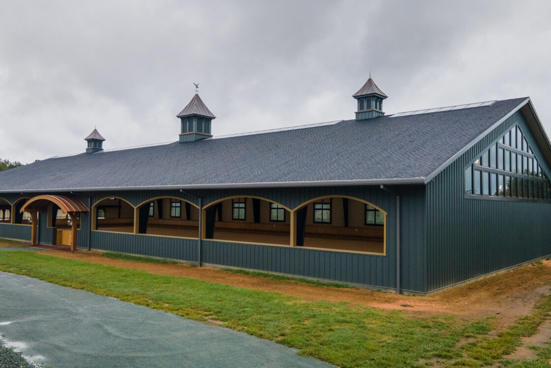 A large, dark gray bank barn with three cupolas on the roof, arched openings along the side, and a covered entrance sits on grass beside a paved path under a cloudy sky.