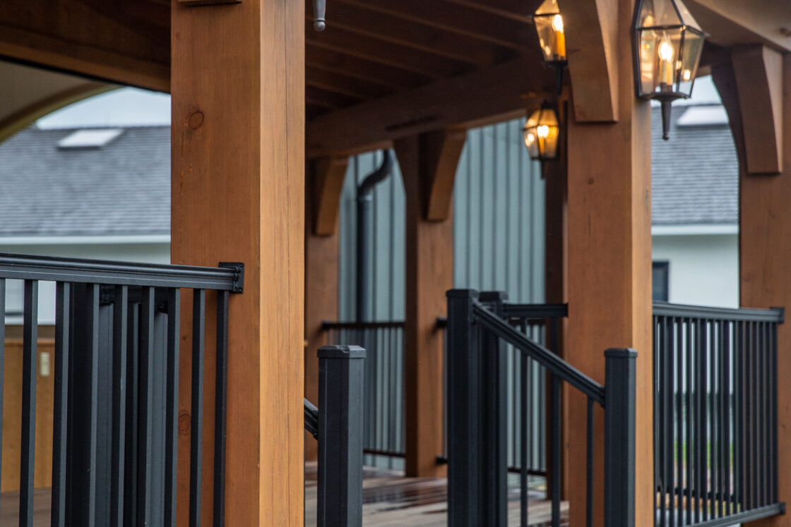 Covered wooden porch with black metal railings, steps leading up, and lantern-style lights hanging from beams. The exposed wooden posts overlook a luxury home styled as a classic bank barn in the background.