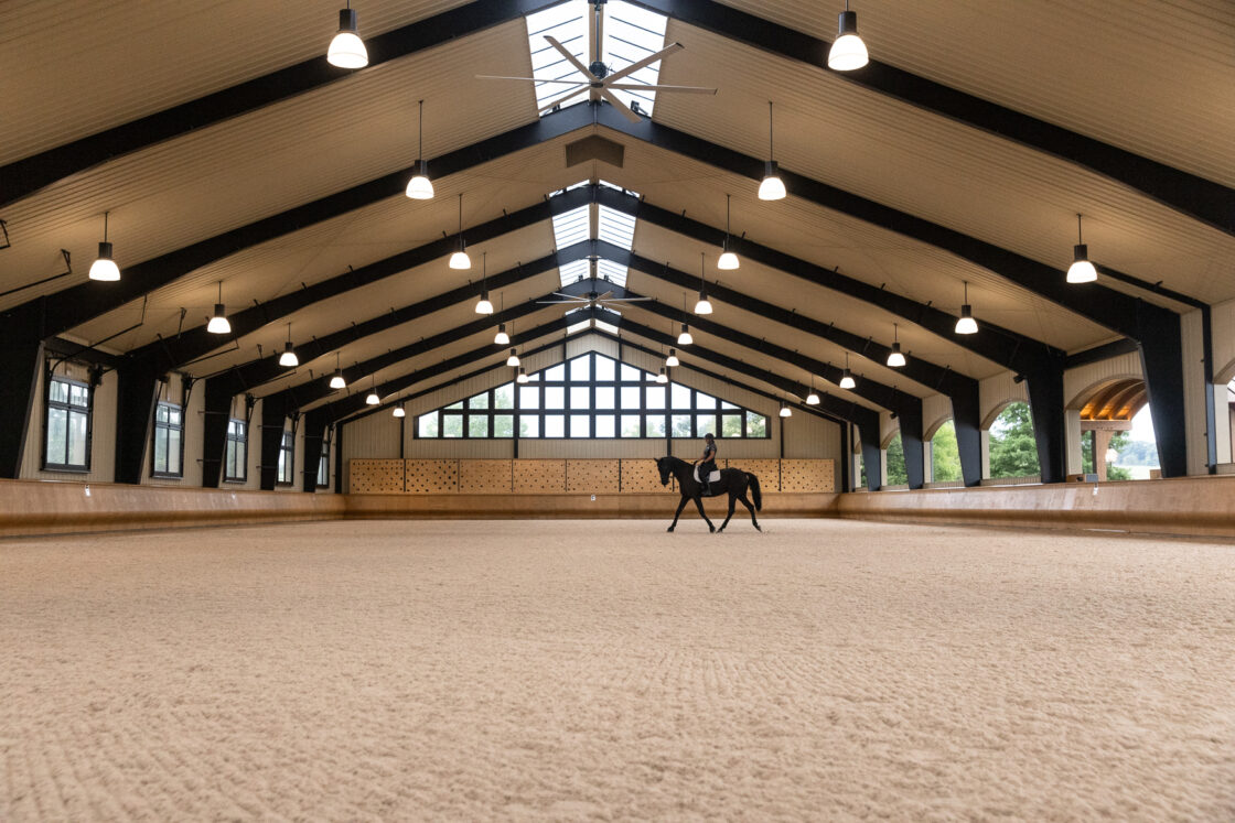 A lone rider on horseback practices in a spacious, well-lit luxury horse barn with large windows, a high vaulted ceiling, and multiple hanging lights.