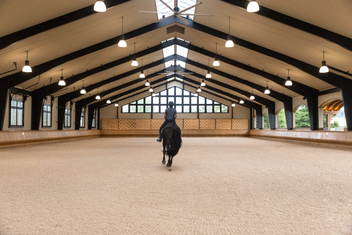 A person rides a black horse inside a spacious, well-lit luxury horse barn arena with large windows, high ceilings, and exposed beams. The view is from behind, facing the far end of the arena.