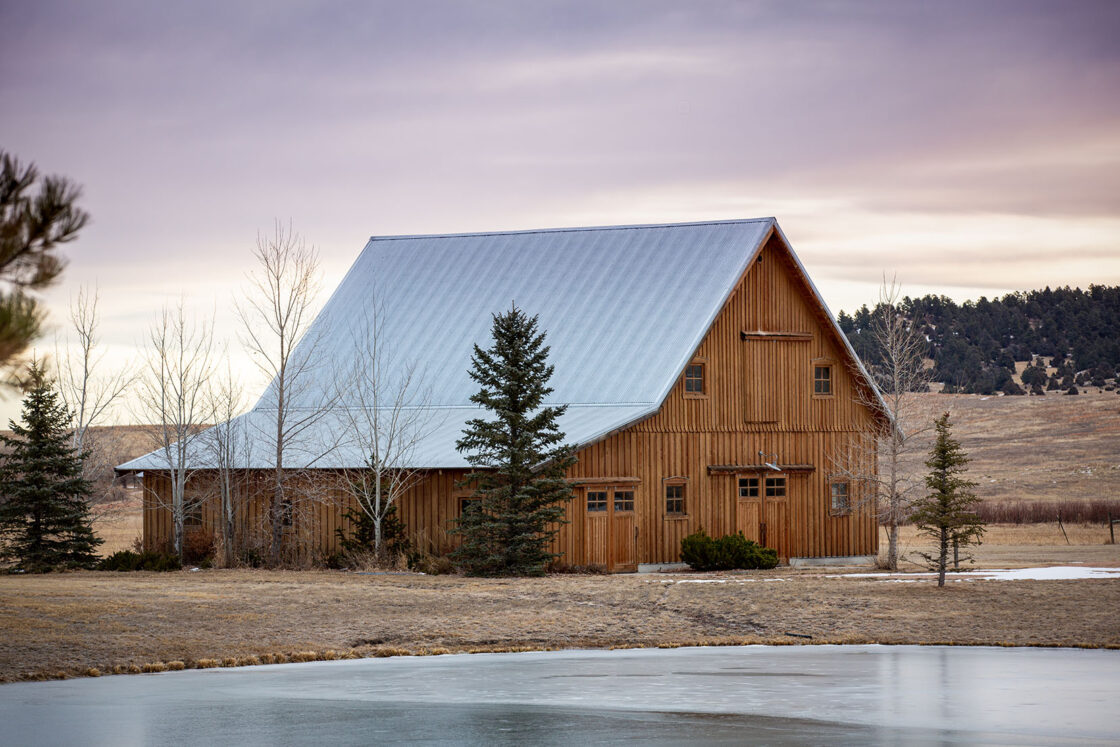 A charming party barn with a metal roof stands near a small pond, surrounded by leafless trees and pines, set in a rural landscape under a cloudy sky at dusk.