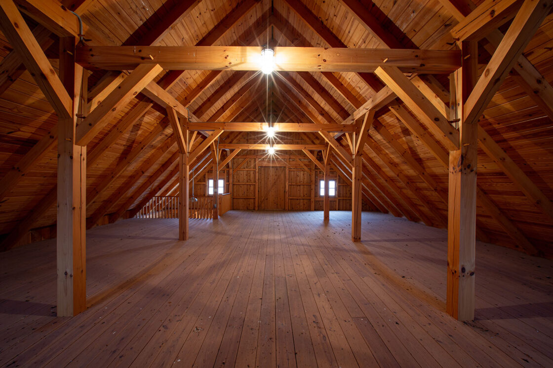 Spacious attic with exposed wooden beams, sloped ceiling, and wooden floor—reminiscent of a luxury home or bank barn; ceiling lights and far-end windows fill the space with natural light.