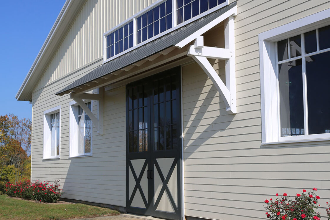 A cream-colored party barn with white trim and large black double doors, surrounded by grass and a few blooming rose bushes, sits under a clear blue sky.