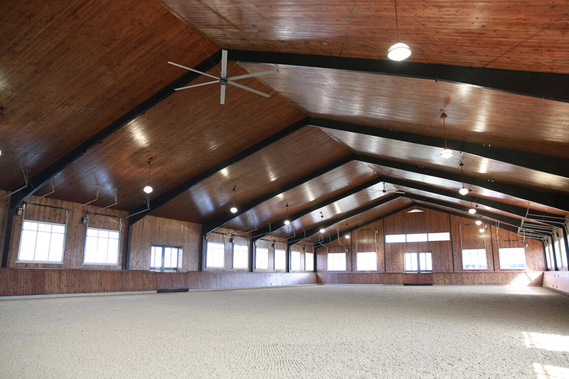 A spacious indoor arena with high wooden ceilings, large windows, bright lighting, and a sand-covered floor—this inviting space combines the charm of a bank barn with modern luxury touches like visible beams and ceiling fans overhead.