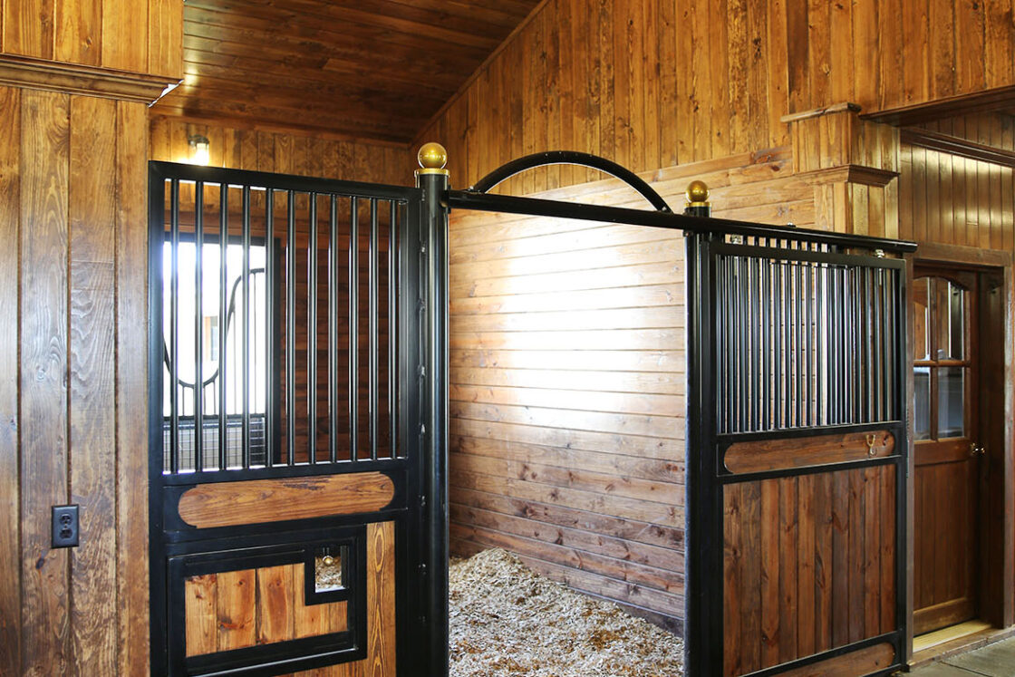 A clean horse stall with wooden walls, a gate featuring metal bars and wood panels, and fresh bedding on the floor, located inside a well-lit, wood-paneled bank barn.