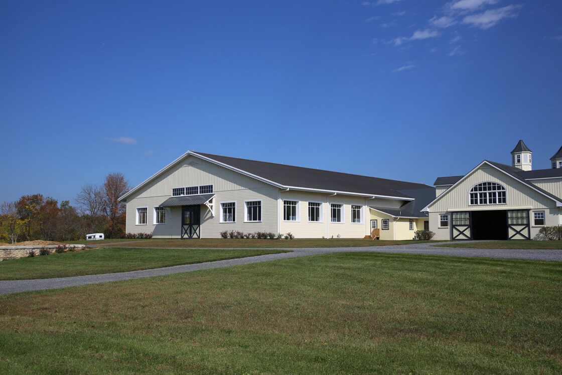 A large light-gray luxury horse barn with white trim and black doors sits on a well-kept green lawn under a clear blue sky. A paved driveway curves in front of the building, and trees are visible in the background.