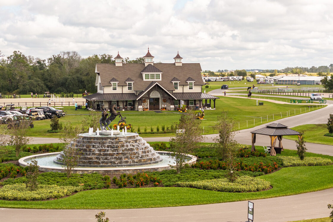 decorative water fountain featuring bronze horse statues that is surrounded by well landscaped green plants which all sit behind a welcome center that has a path to the grounds interior to the right