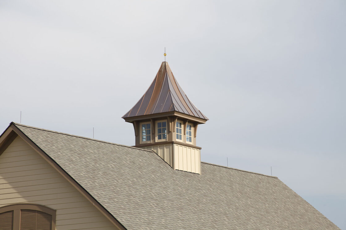A small cupola with windows and a pointed copper roof sits atop a beige luxury horse barn with gray shingles, set against a cloudy sky.