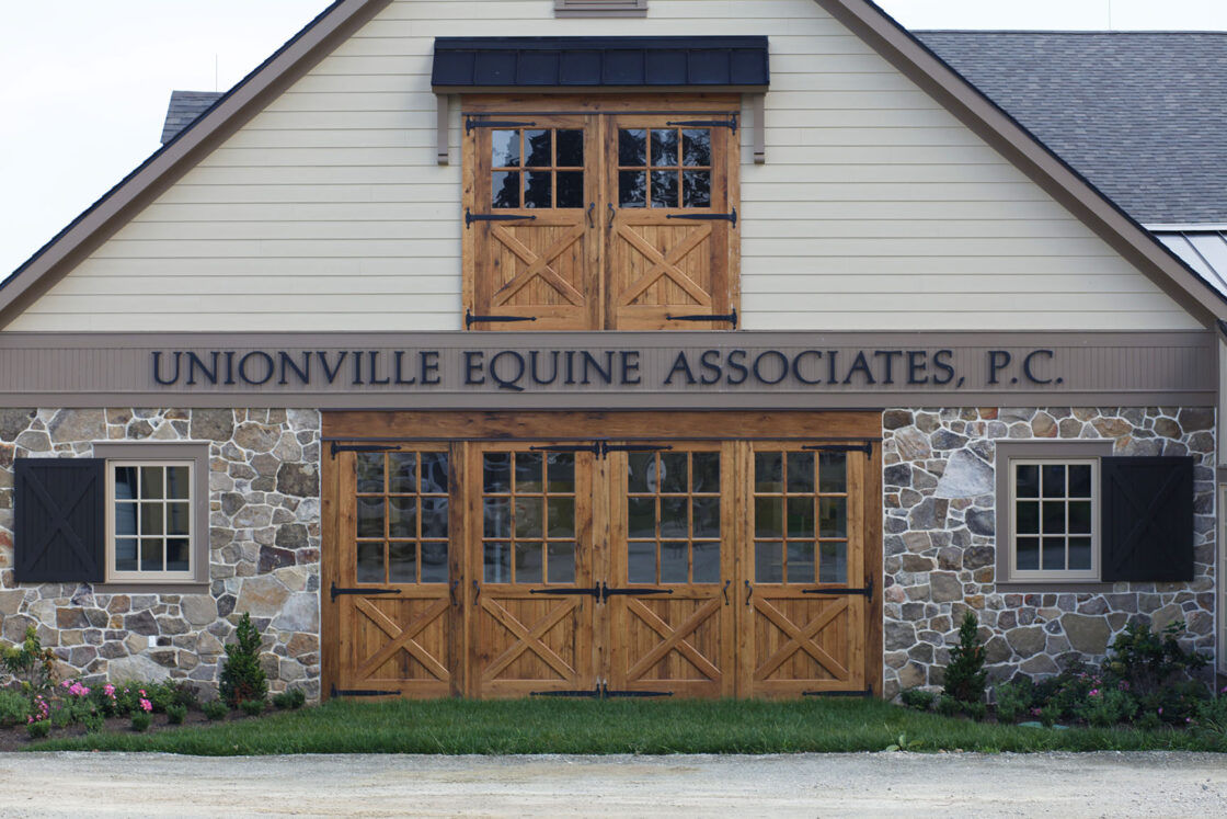 A stone and wood bank barn with large barn doors and the sign Unionville Equine Associates, P.C. above the entrance. Small shrubs and pink flowers line the front.