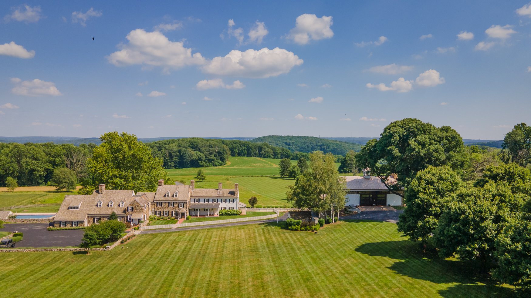 Aerial view of a large country estate with two houses, a luxury horse barn, neatly mowed lawns, tall trees, and expansive green fields under a blue sky with scattered clouds.