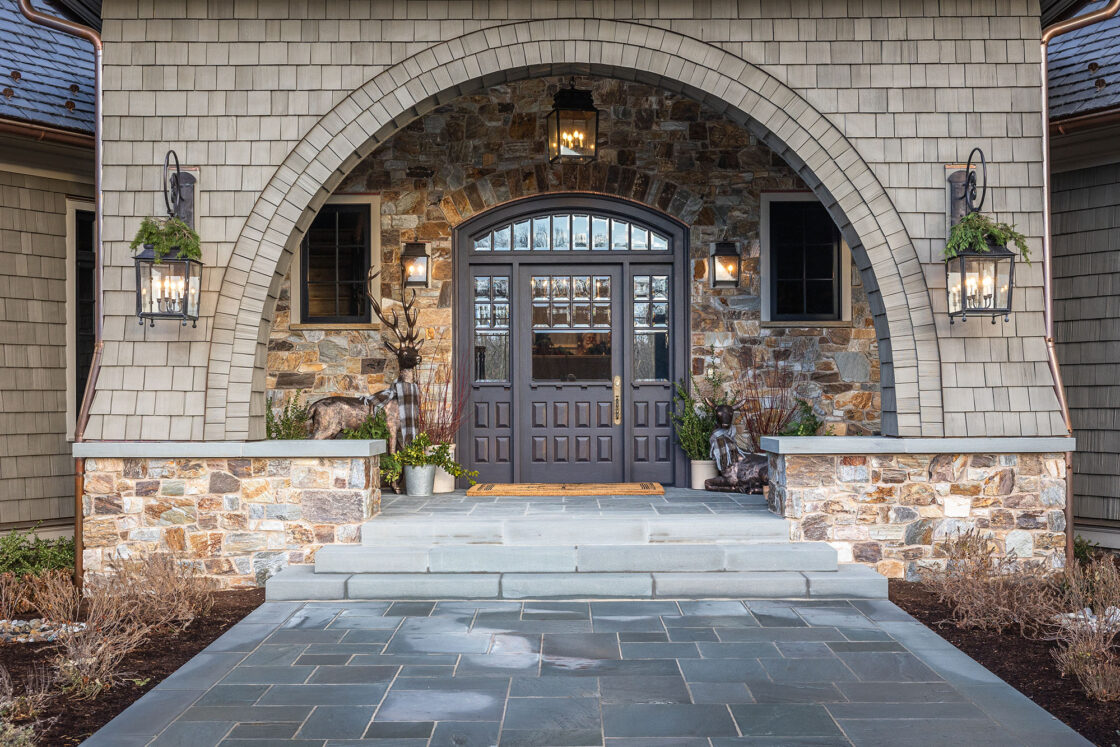 custom wooden doors with large window panels sitting a amidst a luxury home’s stone siding which sits behind a larger archway with a stone walkway leading up to it
