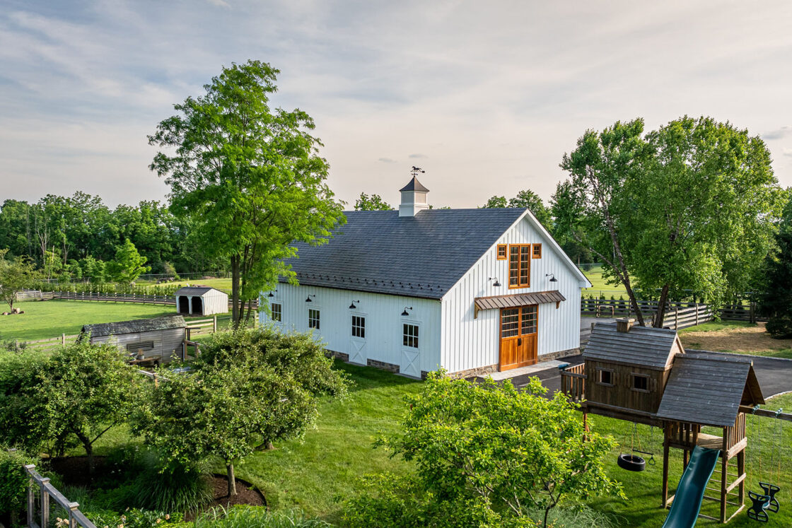 A barn with a swing set and a tree house sitting amidst green trees which sit beneath a clear sky