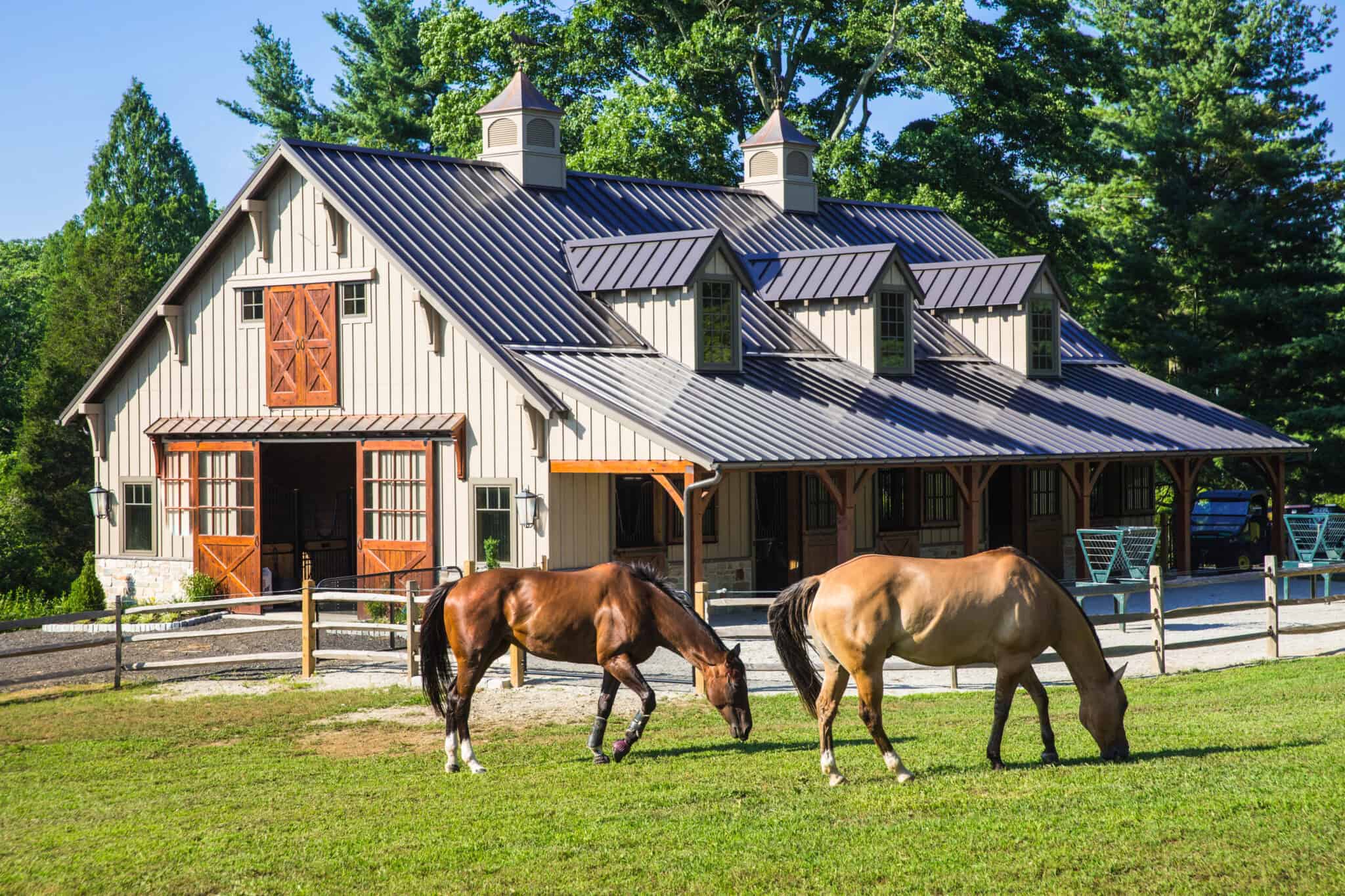 Two horses graze on green grass in front of a luxury horse barn with dormer windows, wooden doors, and a metal roof, surrounded by trees and a wooden fence on a sunny day.