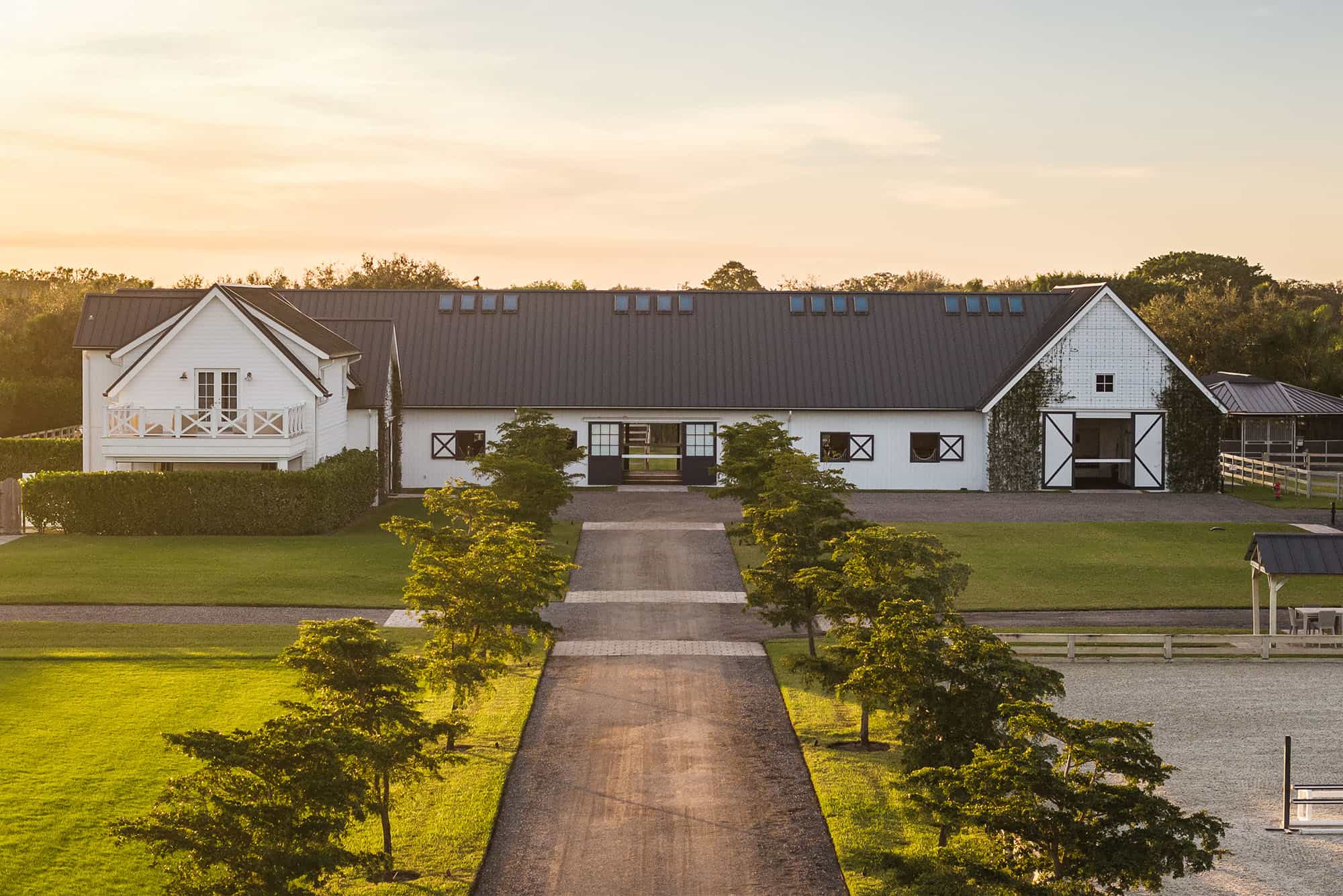 A wide gravel driveway lined with trees leads to a luxury horse barn featuring black accents and a dark metal roof, surrounded by green lawns and fencing under a clear sky at sunset.