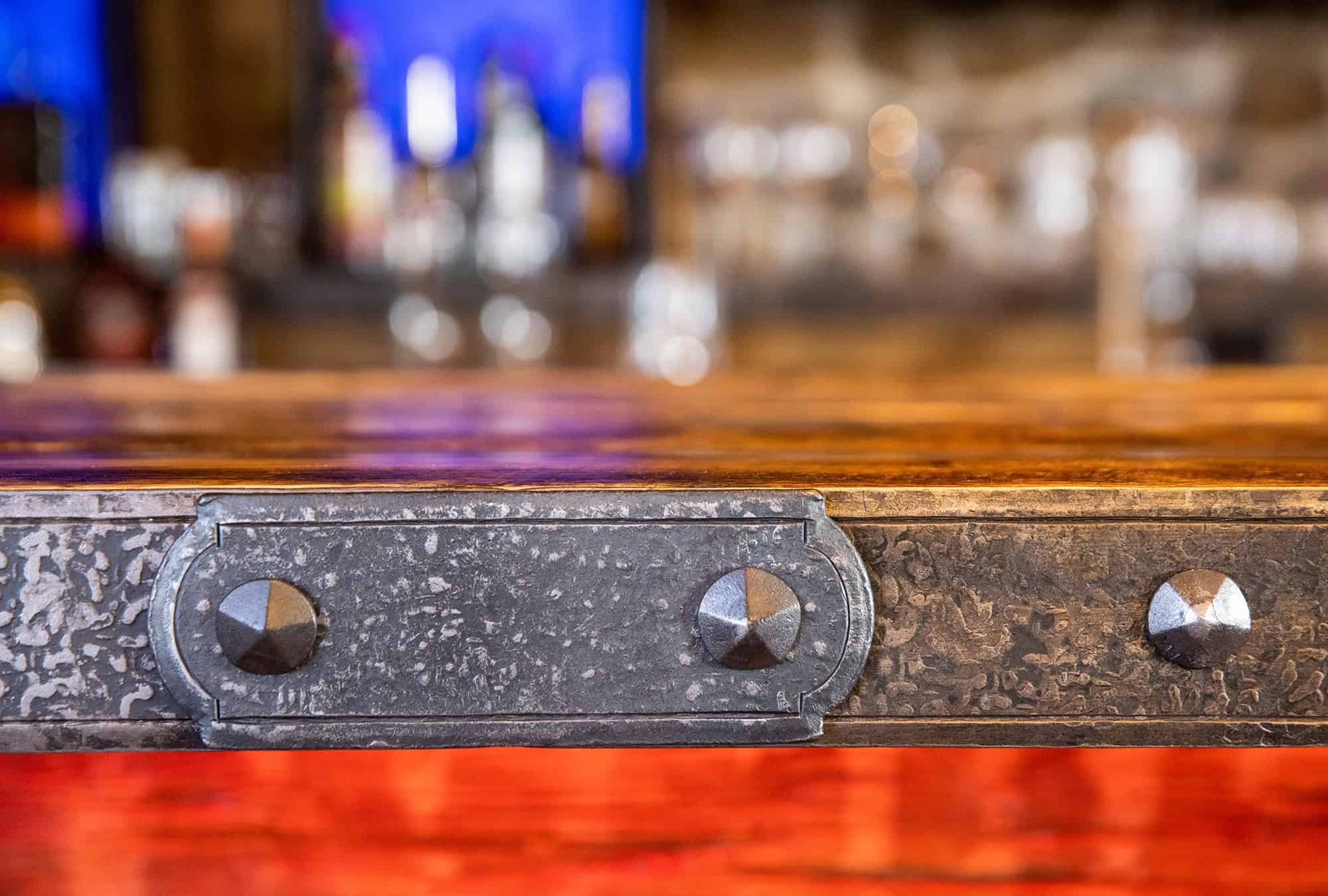 Close-up of a rustic, metal-edged wooden tabletop with visible bolts. The blurred background, reminiscent of a luxury horse barn or bank barn, features colorful lights and indistinct shapes, evoking a warm bar or restaurant setting.