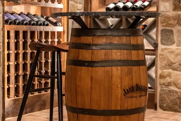 A wooden barrel with a tabletop and two wooden stools sits in a wine cellar, surrounded by stone walls, shelves, and racks filled with wine bottles—creating an ambiance reminiscent of a luxury horse barn.