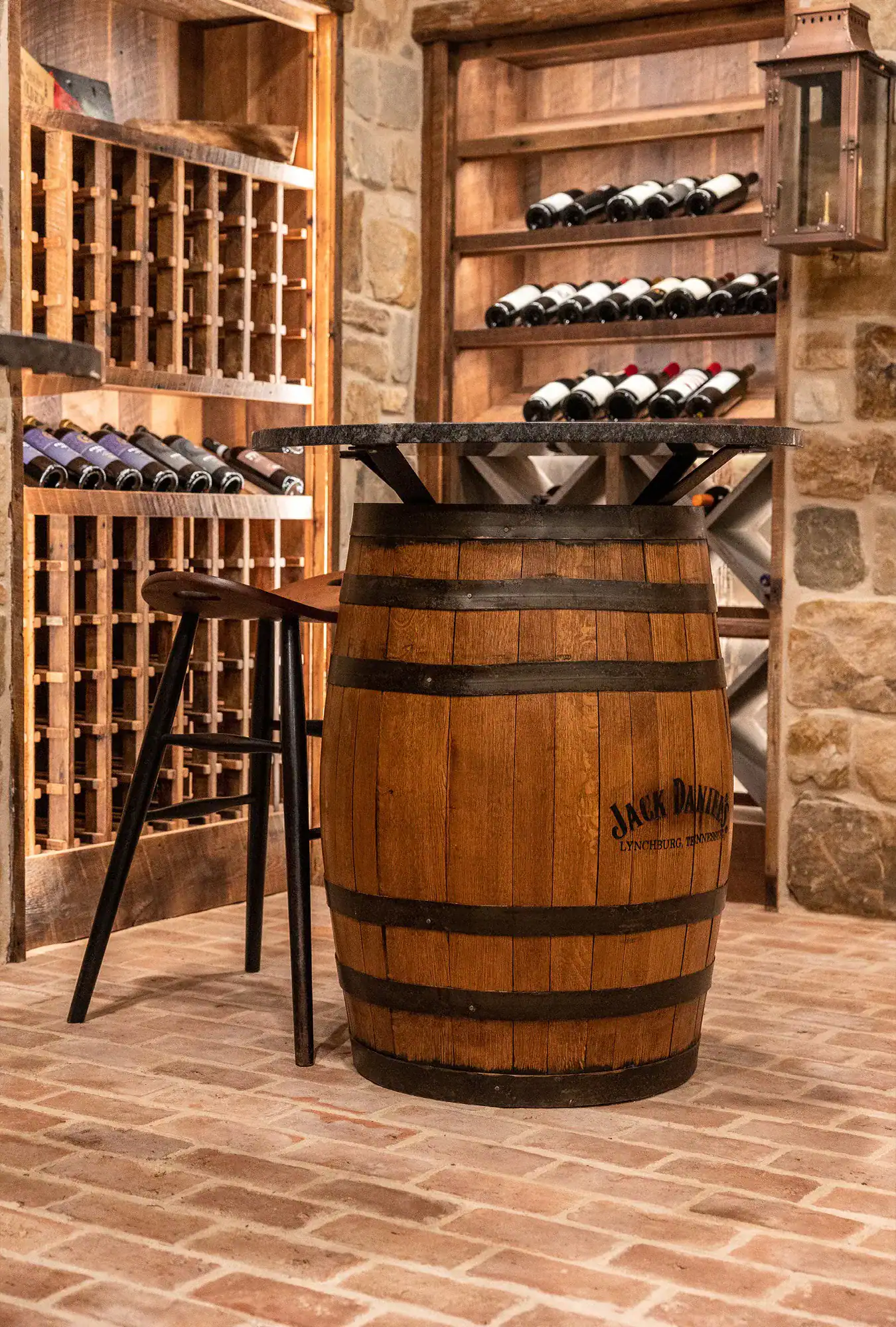 A wooden barrel with a tabletop and two wooden stools sits in a wine cellar, surrounded by stone walls, shelves, and racks filled with wine bottles—creating an ambiance reminiscent of a luxury horse barn.