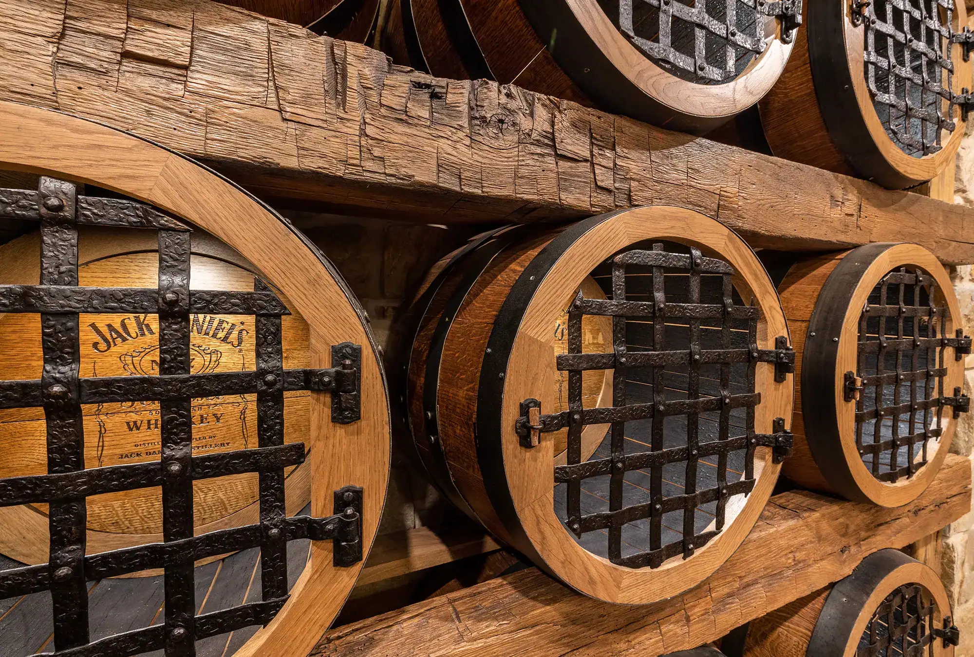 Wooden whiskey barrels with metal grates are stacked on wooden shelves. The side of one barrel is marked with the Jack Daniel’s whiskey logo. The scene evokes a rustic storage area perfect for a luxury horse barn or party barn setting.