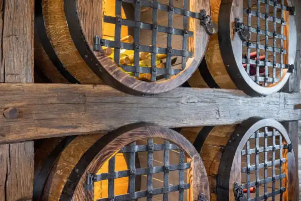 Large wooden barrels with metal grates are built into a rustic wooden wall, storing and displaying wine bottles inside a warmly lit wine cellar reminiscent of a luxury home or party barn.